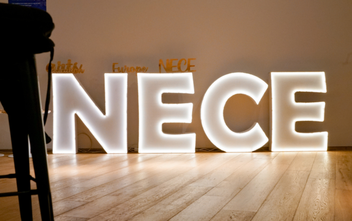 Illuminated letters spelling "NECE" against a wall on a wooden floor.